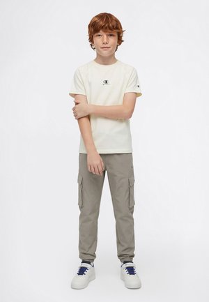 Jongen met rood haar die een wit Champion T-shirt, grijze cargobroek en witte sneakers met blauwe veters draagt, staand tegen een effen achtergrond.