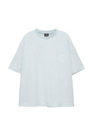 Junge Person mit lockigem Haar, die ein hellblaues T-Shirt mit kleinem weißem "Grande Squadra"-Logo auf der Brust trägt.
