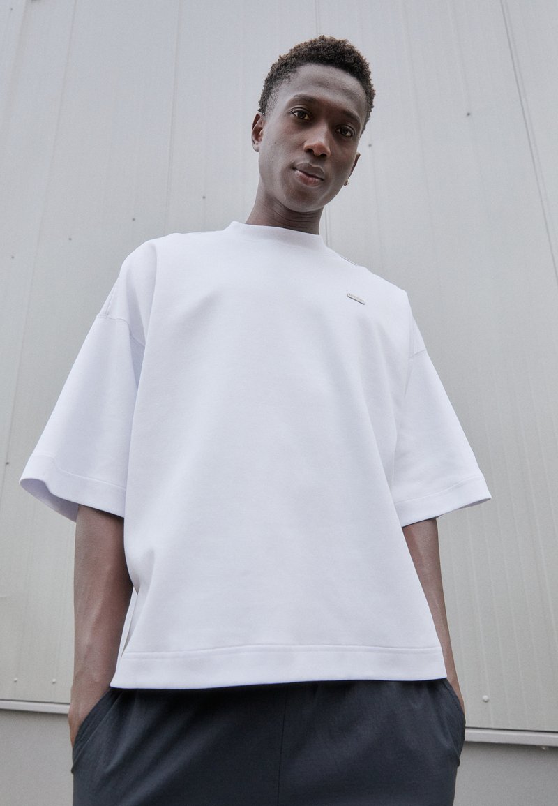 Hvit oversized t-skjorte med korte ermer, som har rund hals og subtile logo aksenter. Stoffet ser mykt ut med en glatt tekstur.