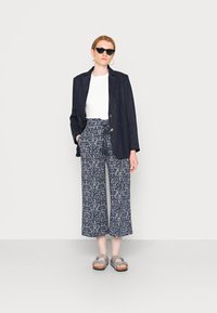 Blazer de linho azul marinho, camisa branca ajustada, calças largas estampadas, sandálias cinzentas. O blazer tem dois botões; as calças têm um cós com cordão.