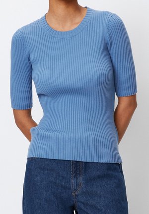 Femme portant un pull ajusté côtelé à manches courtes bleu clair associé à un jean bleu foncé sur un fond uni.