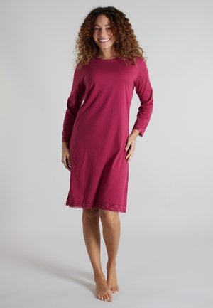 Smiling woman with curly hair wearing a long-sleeve, knee-length dark pink dress with lace trim, standing barefoot on a light background.