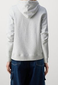 Felpa grigia chiaro con cappuccio, vestibilità comoda, polsini a costine e tessuto liscio. Abbinata a jeans blu larghi.