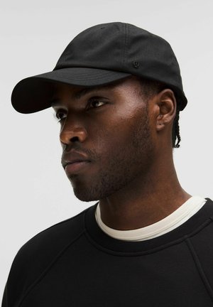 Černá čepice s prohnutým kšiltem, vyrobená z měkké tkaniny. Má minimalistické značení a je kombinována s tmavou mikinou a bílým límečkem.