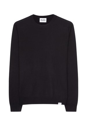 Giovane uomo con capelli corti castani che indossa un maglione nero con collo rotondo e pantaloni blu navy, mani nelle tasche, che guarda in basso.