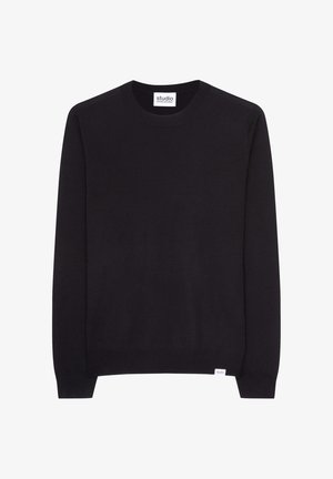 Ung mand med kort brunt hår iført en sort crewneck-trøje og marineblå bukser, med hænderne i lommerne og blikket rettet nedad.