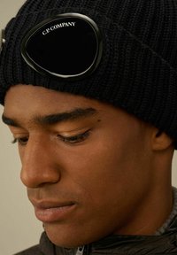 Berretto di maglia nera con un accessorio per occhiali da sci dalla tonalità verde prominente. Primo piano del viso, che mette in mostra la texture della pelle e le sottili caratteristiche facciali.