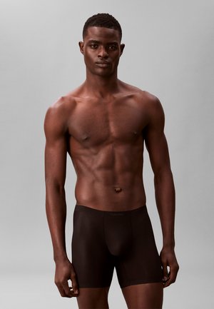 Atletyczny mężczyzna stoi ubrany w czarne bokserki Calvin Klein na jednolitym szarym tle.