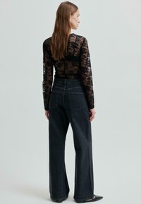 Kobieta z długimi brązowymi włosami, ubrana w czarną koronkową bluzkę z długim rękawem oraz czarne dżinsy z wysokim stanem o szerokich nogawkach, stojąca tyłem.