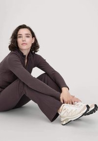 Bruine geribbelde sportset met een hoge kraag, gecombineerd met witte en beige sneakers met een gestructureerde zool. Neutrale achtergrond.
