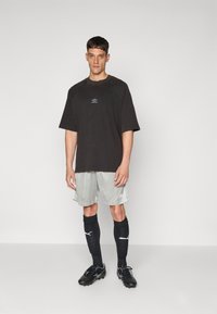 Černé oversized tričko s logem, světle šedé šortky, černé fotbalové ponožky; černé kopačky s bílými detaily; jednoduchý, sportovní design.