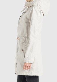 Giacca impermeabile beige chiaro con cappuccio, vita regolabile con coulisse, chiusura con zip e tasche con bottone a pressione. Texture liscia e design elegante.