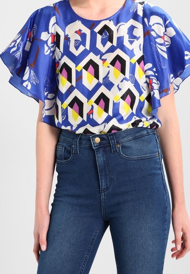 Kortærmet, blå bluse med mønster af geometriske former i gul, sort og hvid. Sammen med højtaljede, mørkeblå jeans.
