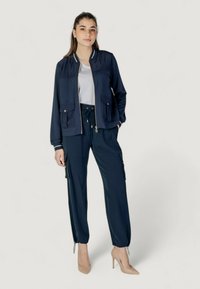 Giacca bombardiere blu navy con due tasche frontali, abbinata a pantaloni cargo blu navy. Entrambi i capi presentano una texture liscia e dettagli di design funzionali.