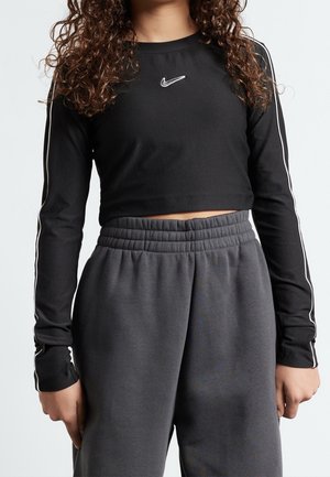 Personne portant un crop top noir à manches longues Nike avec des rayures blanches sur les manches et un pantalon gris foncé à taille élastique.