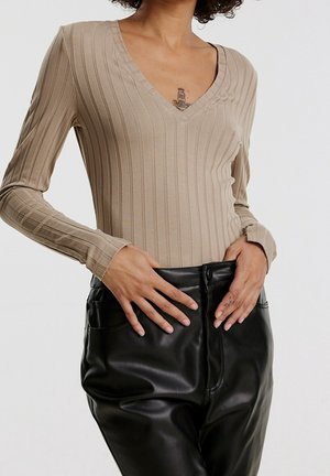 Femme portant un haut beige côtelé à col en V manches longues et un pantalon en cuir noir taille haute, les mains reposant près de la taille.
