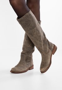 Bottes en cuir marron montant jusqu'au genou avec un motif texturé, des bouts arrondis et un petit talon empilé, présentant une finition usée et des coutures visibles.