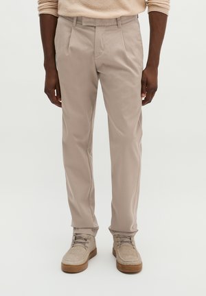 Mann trägt beige Straight-Fit-Hosen und passende beige Schnürschuhe, steht vor neutralem Hintergrund.