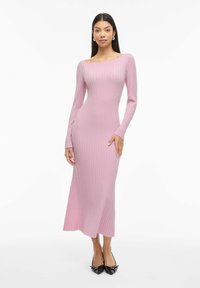 Moteris dėvi prigludusią, ilgomis rankovėmis, rausvą ribanuotą midi ilgio suknelę ir juodus aštriais galiukais aukštakulnius, stovėdama prieš baltą foną.