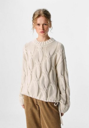 Donna che indossa un maglione cream oversize a treccia e pantaloni di velluto a coste marroni, in piedi contro uno sfondo bianco puro.
