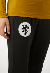 Svarte treningsbukser har en sirkulær hvit lapp med et svart løveemblem. Materialet er glatt, med en tettsittende design og sidelommer.