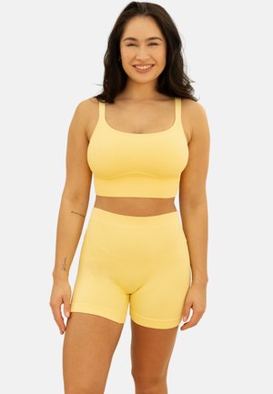 Lächelnde Frau mit langen dunklen Haaren, die einen passenden, gerippten gelben Crop-Top und eine hoch taillierte Shorts trägt, steht vor einem schlichten Hintergrund.