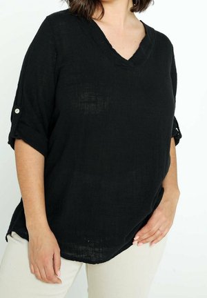 Femme portant un haut noir ample à col en V avec des manches retroussées et un pantalon blanc, posant avec une main posée sur la hanche et l'autre sur le côté.