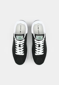 Baskets noires en toile avec semelles en caoutchouc blanc et lacets, présentant des étiquettes de langue de marque vertes et blanches et des accents de couture ton sur ton.