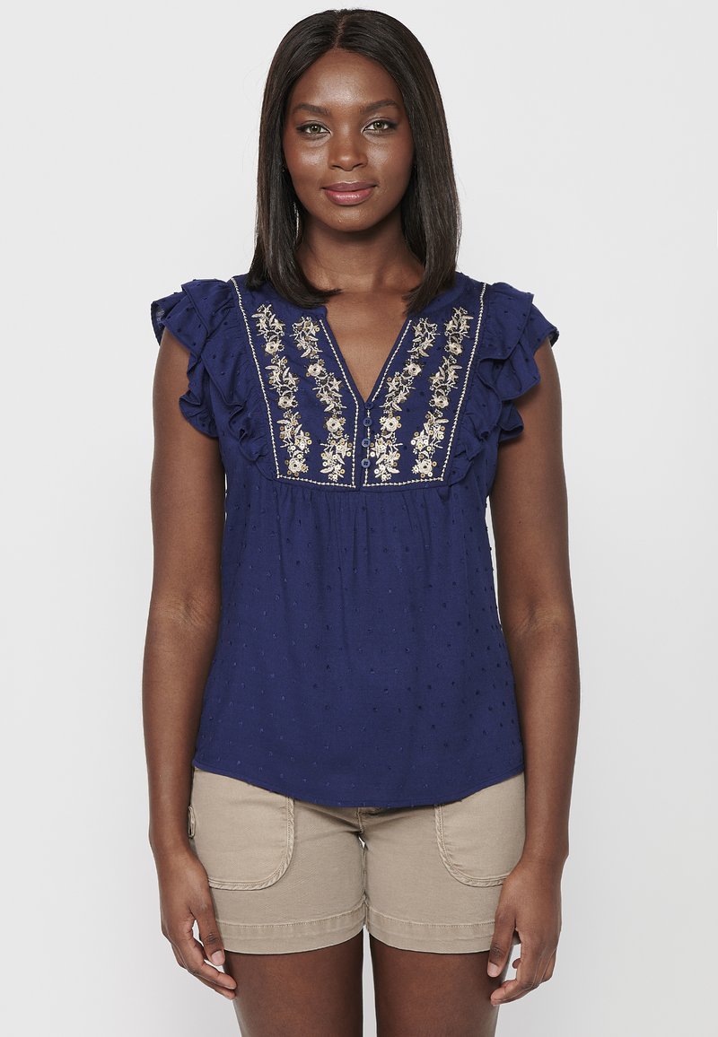 Marinebluse mit kurzen Rüschensäumen, verziert mit goldener Blumenstickerei entlang des Ausschnitts und gepunkteter Textur. Kombiniert mit beigen Shorts.