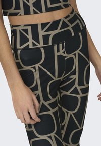 Zwarte leggings met een geometrisch beige patroon, gemaakt van rekbaar materiaal. De tailleband is elastisch met een vlakke naadontwerp.