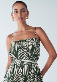 Zaļas krāsas rakstains kleita ar baltu lapu motīviem, ar plānām lencītēm un savilktu kakla daļu, jostasvietā satīta ar zelta gredzena akcents.