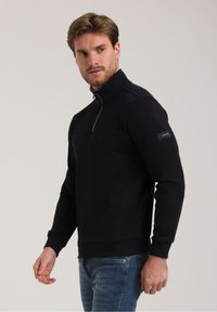 Schwarze Zip-up-Sweatjacke mit strukturiertem Einsatz über der Brust. Mit gerippten Bündchen und Saum sowie einem Logopatch am Ärmel.