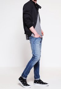 Schwarze Bomberjacke, graues T-Shirt, hellblaue Jeans und schwarze Sneakers mit weißen Mustern; Modell steht im Profil.