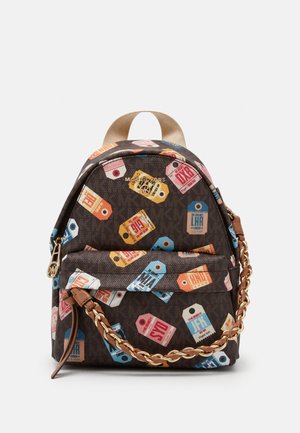Petit sac à dos marron avec des motifs colorés d’étiquettes de voyage, bandoulière chaîne dorée, poche avant zippée et poignée supérieure beige.