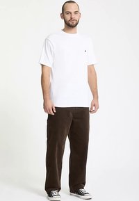 Maglietta bianca in cotone con maniche corte e piccolo logo sul petto, indossata con pantaloni in velluto marrone e scarpe da ginnastica nere con lacci bianchi.