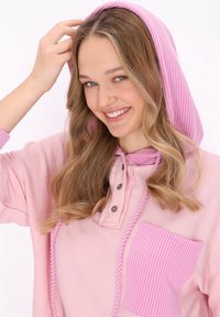 Růžová mikina s pruhovanou kapucí, knoflíkovou légou a texturovanou přední kapsou. Měkká tkanina a volný střih.
