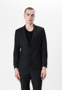 Zwarte colbert met een notch-lapel, enkelvoudig ontwerp, twee knopen en zakdetails; gedragen met een zwart hemd eronder.