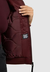Bordeaux gewatteerde jas met een interieur met geribde tailleband, ritsluiting en een logopatch met zwarte tekst op een witte achtergrond.