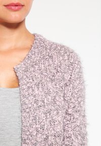 Stickad kofta i ljusrosa med texturerat garn. Har rund hals och öppen frontdesign, tillsammans med en grå topp under.