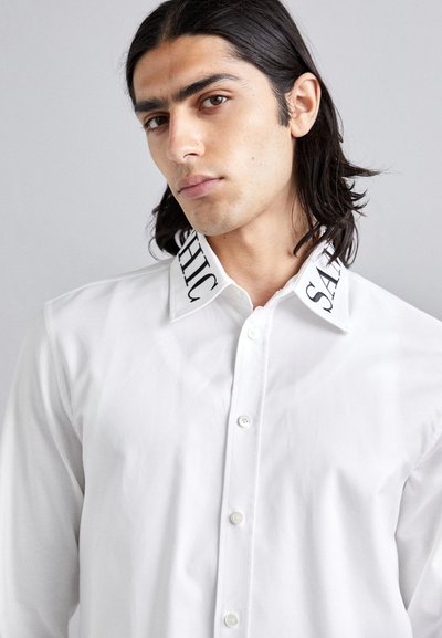 White button-up shirt with a smooth texture, featuring a contrasting black print on the collar that reads "CHIC" and "SACH."