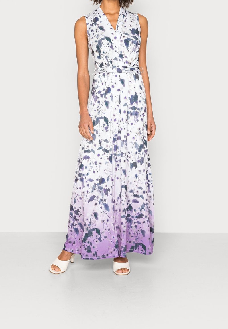 Robe maxi sans manches à col en V avec un motif floral blanc, noir et violet, taille cintrée par une ceinture, portée avec des talons ouverts blancs.