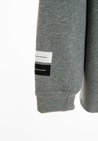 Manga de sudadera gris con puño acanalado que presenta dos etiquetas de tela; una blanca y una negra, ambas con texto impreso. Textura suave.