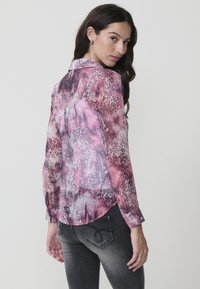 Blusa transparente de manga larga en estampado animal en tonos rosa, morado y gris, con cuello y dobladillo curvado, acompañada de jeans gris oscuro.