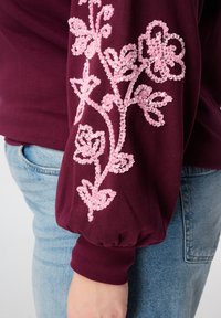 Bordeauxrode sweatshirt met ballonmouwen, versierd met roze bloemrijke borduursels en kralenaccenten op de mouwen. Spijkerbroek is zichtbaar.