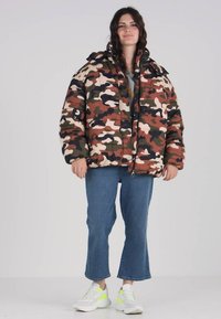 Maskuojantis pūkinis švarkas ramiai žemės spalvų gama, su aukštu apykakle, priekyje užtrauktuku ir horizontaliai siuvinėtu raštu. Sujungtas su mėlynomis džinsomis ir baltomis sportbačiais.