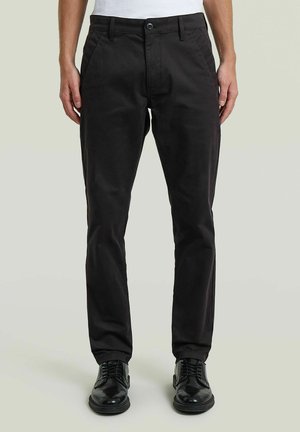 Homme portant un pantalon noir slim-fit avec poches latérales, fermeture par bouton et zip, associé à des chaussures de ville noires et brillantes.
