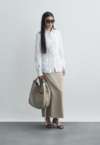 Balta krekla ar teksturētu priekšpusi, bēša garā svārki, melnas sandalēs un lielā bēšā somā ar zelta logo akcentiem. Gludās tekstūras un modernas dizains.