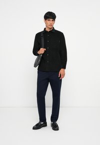 Schwarzes Cordhemd mit Knopfverschluss, einer Brusttasche, kombiniert mit dunkelblauen Hosen und schwarzen Slippern, mit einer kleinen grauen Tasche in der Hand.