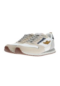 Sneaker beige e bianche con lacci grigi, materiali in camoscio e rete, suola in gomma con pattern maculato e emblemma dorato con ali sul lato.