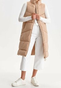 Gepolsterte, ärmellose Weste in hellbeige mit hohem Kragen. Hat einen Reißverschluss vorne und Seitentaschen. Wird über einem weißen Langarmshirt und einer weißen Hose getragen.